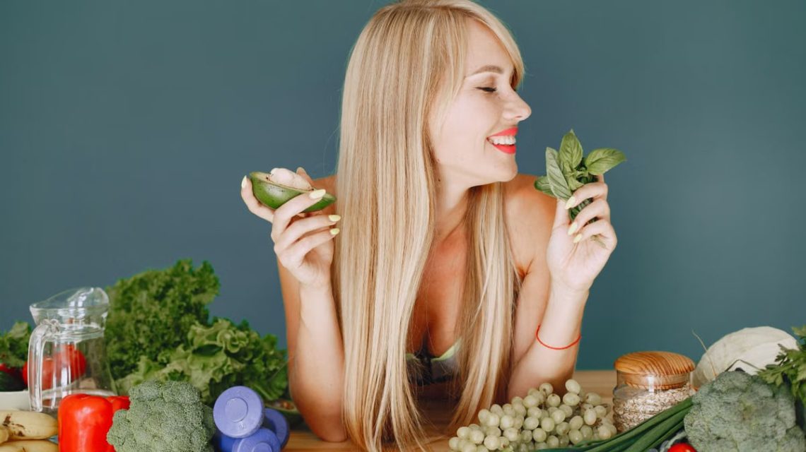 A smiling woman holding avocado and basil, surrounded by fresh vegetables and fruits for healthy eating.